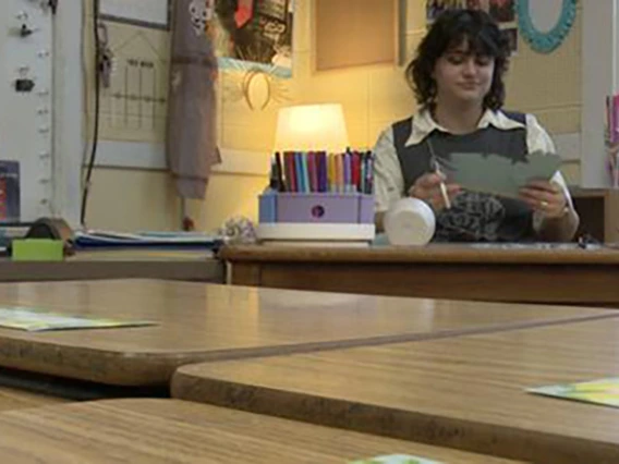 inez mcclain sitting at a teachers desk in a classroom with a lamp lit behind her