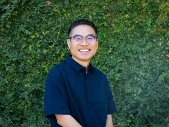 ahn headshot, wearing a blue shirt, eyeglasses, greenery in the background