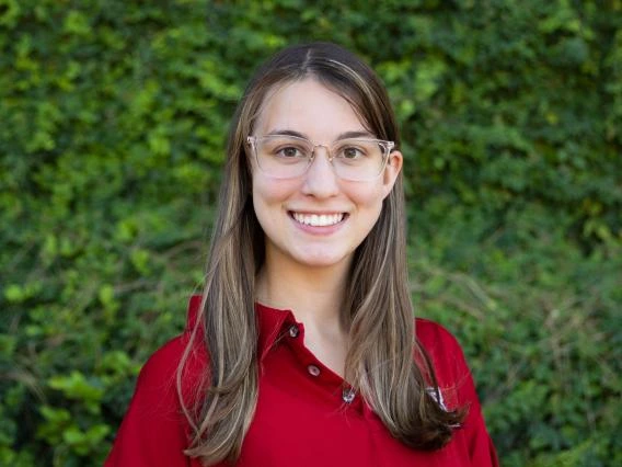 Mackenzie Frithsen wearing a red shirt, eye glasses with greenery in the background