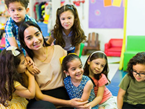 young educator surrounded by young children hugging her in a classroom setting