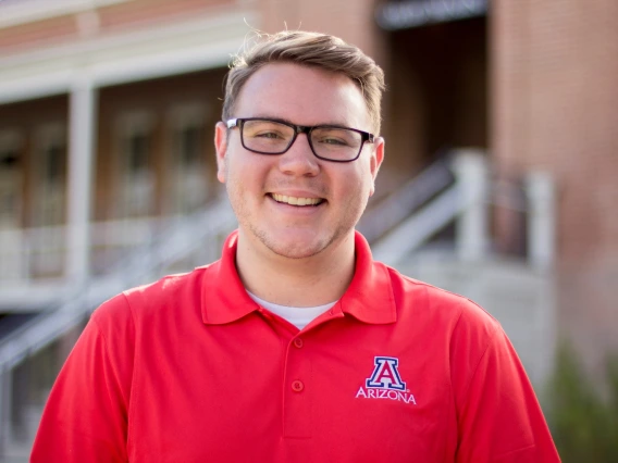 Kyle Fisher in a red shirt, wearing eye glasses, outside in front of old main building