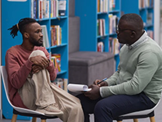 two men sitting across from each other talking, books on shelves in the distance