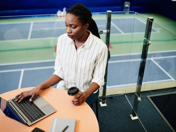 woman sitting at a table with laptop, tennis court as a backdrop