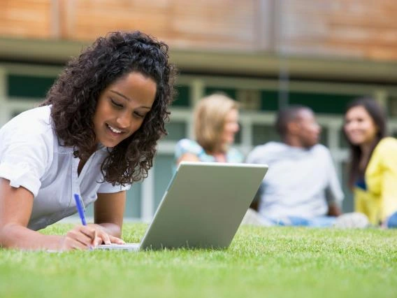 students sitting on the grass, one working on a computer, the others talking in the distance