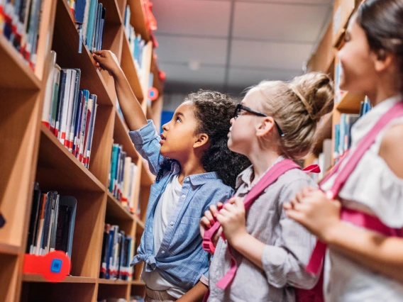 three grade school kids in a library aisle looking at books