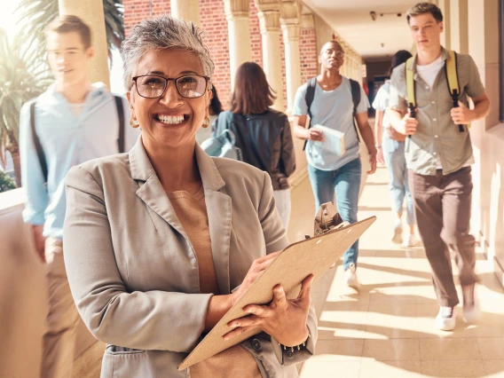 woman wearing glasses holding a clipboard and students in the distance walking with backpacks