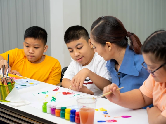 young teacher sitting with three students at a table painting