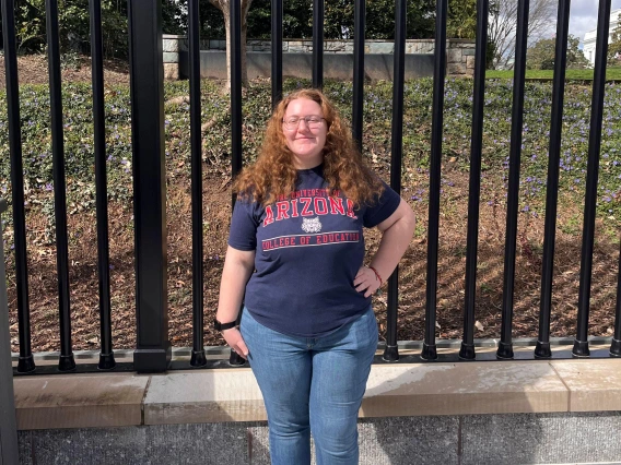 christina petrin standing outdoors under a sunny sky, black fence and greenery in the background