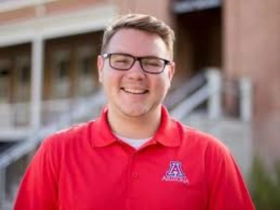 kyle fisher headshot, wearing a red UA polo and eye glasses, old main in the background