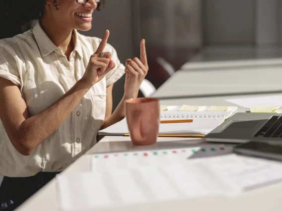 Smiling young lady learning and communicating in sign language online while sitting at workplace