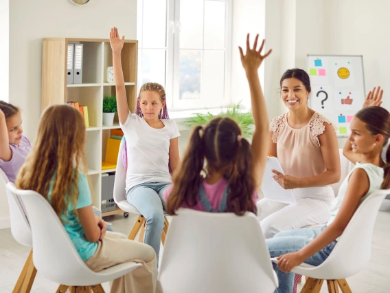 Group of school children girls sitting in a circle with female friendly woman psychologist and raising their hands up to ask or answer a question during a meeting on therapy session