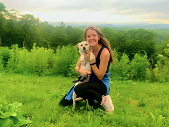 Alison Irwin kneeling in a grassy field, holding a small dog, sunset in the distance