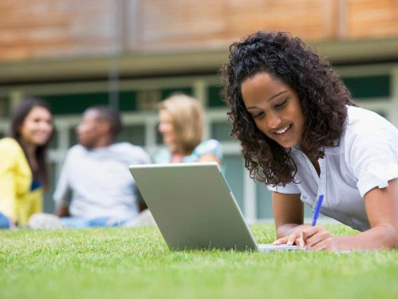 students sitting on the grass, one working on a computer, the others talking in the distance