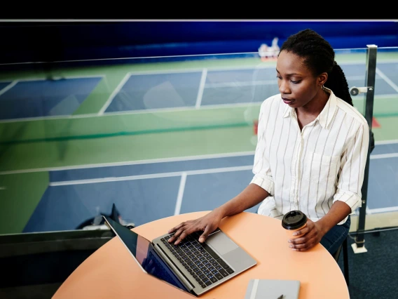 woman sitting at a table with laptop, tennis court as a backdrop