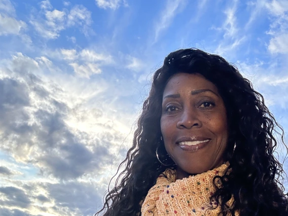 A woman with long, curly hair wearing a beige knitted sweater with colorful speckles, smiling against a scenic backdrop of a blue sky with wispy clouds and a golden sunset.