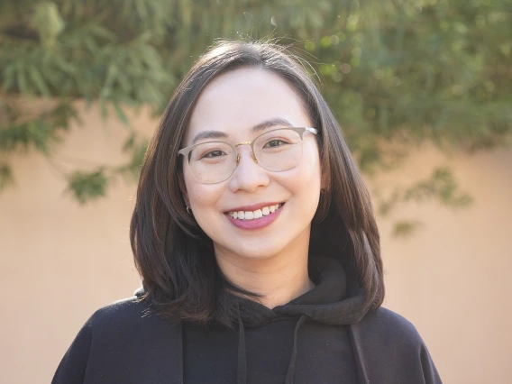 A woman with shoulder-length dark hair and glasses, smiling while wearing a black hoodie with an embroidered design, standing outdoors with green foliage in the background.