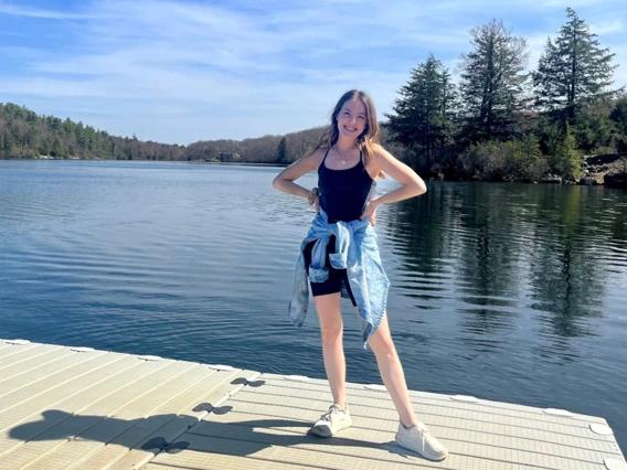 A young woman with long, wavy brown hair stands on a floating dock, smiling with her hands on her hips. She is wearing a black tank top and shorts, with a light blue denim jacket tied around her waist and white sneakers. The background features a calm lake surrounded by trees under a bright blue sky.