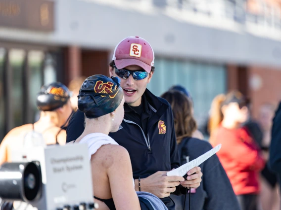 A swim coach wearing a red USC cap, sunglasses, and a black jacket with the USC Trojans logo speaks with a swimmer wearing a black swim cap with the USC logo and a white towel draped over their shoulders. The coach is holding a piece of paper and appears to be giving instructions. Other swimmers and team members are visible in the background at the poolside.