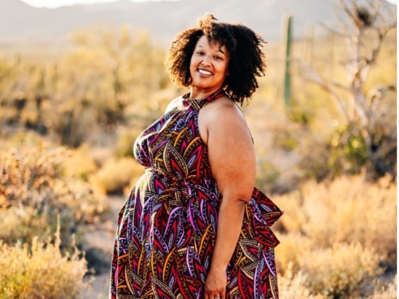 A smiling woman with curly black hair stands in a desert landscape during golden hour. She is wearing a colorful, patterned maxi dress with a high neckline. The background features cacti, desert shrubs, and distant mountains bathed in warm sunlight.