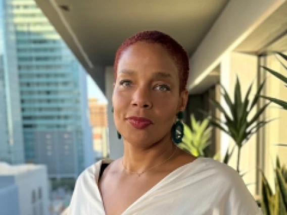 Portrait of a woman with short red hair, wearing a white blouse and dark earrings. She stands on a balcony with a cityscape of modern buildings and greenery in the background, looking confidently at the camera.