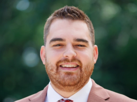 Portrait of a smiling man with short brown hair and a beard, wearing a rust-colored suit, white dress shirt, and a red and white patterned tie. He is standing outdoors with a blurred green foliage background.