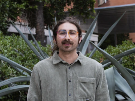 headshot of a Latino man with long hair tied back, wearing rainbow glasses with a septum piercing. He is dressed in a light green, corduroy-style button-up shirt and is standing in front of a large agave plant.     2 / 2   100%