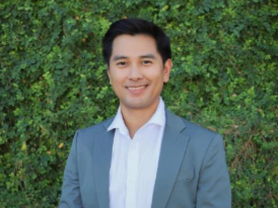 smiling Filipino man standing in front of a lush green wall covered in small, leafy vines. He has short, black hair and is wearing a light gray suit jacket over a white collared shirt. 