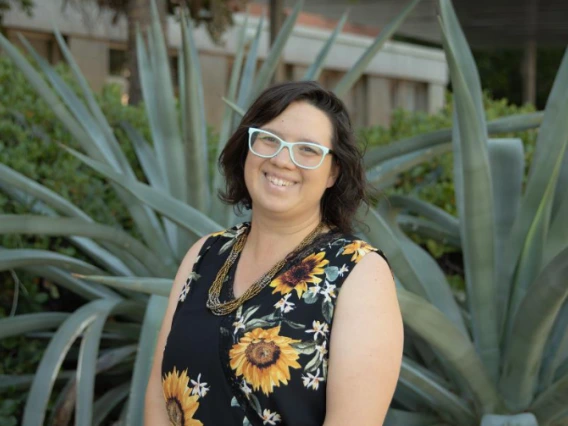 A smiling woman with blue glasses, a brown necklace, and a black top featuring sunflowers, standing in front of an aloe vera plant.  