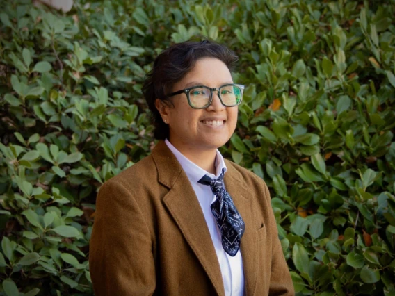 Portrait of a person with short dark hair, wearing glasses, a brown blazer, a white shirt, and a patterned black tie. They are smiling and standing outdoors in front of lush green foliage.