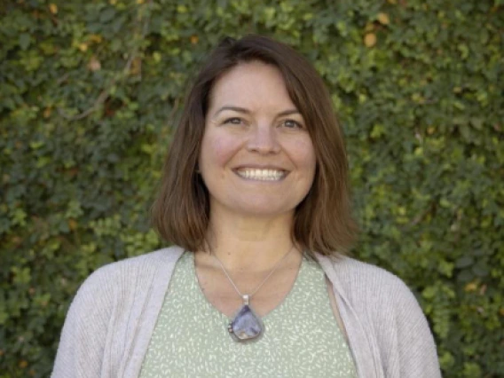 A woman with short brownish hair and brown eyes wears light green and cream vine pattern top, a big purple stone pendant, and a cream shawl. She stands smiling in front of a vine covered wall.