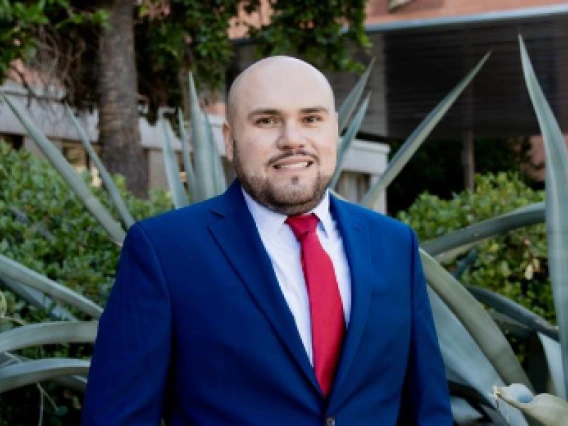 Portrait of a bald man with a beard, wearing a navy blue suit, white shirt, and red tie, smiling at the camera. He is standing outdoors in front of large agave plants and greenery, with a modern building in the background.