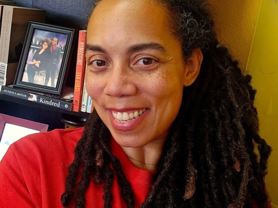 A smiling woman with long, dark dreadlocks wearing a red University of Arizona shirt takes a selfie in front of a bookshelf filled with books, awards, and a framed photograph