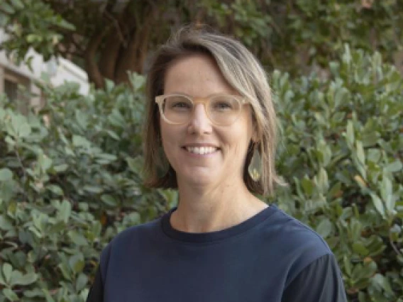 white, middle-aged woman with chin-length light brown hair wearing glasses and standing against a green leafy background