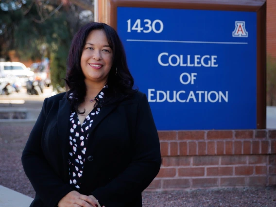 Portrait of a smiling woman with shoulder-length dark hair, wearing a black blazer over a patterned blouse, sitting in front of a blue sign that reads '1430 College of Education' with the University of Arizona logo. The background features a brick building, leafless trees, and parked motorcycles.