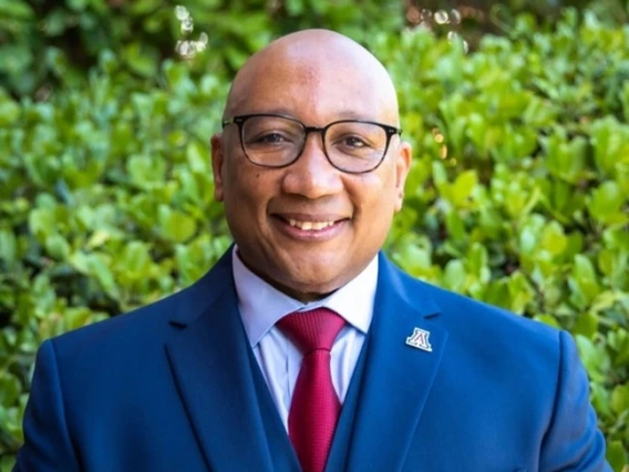 Portrait of Robert Berry, a bald man wearing glasses, a navy blue suit, white dress shirt, and a red tie. He is smiling and standing outdoors in front of green foliage, with a University of Arizona lapel pin on his suit jacket.