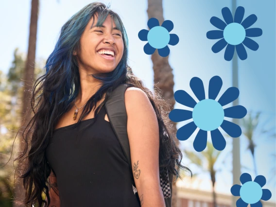 A smiling young woman with long dark hair, blue highlights, and tattoos on her arms walks outside under palm trees, carrying a backpack with a water bottle attached