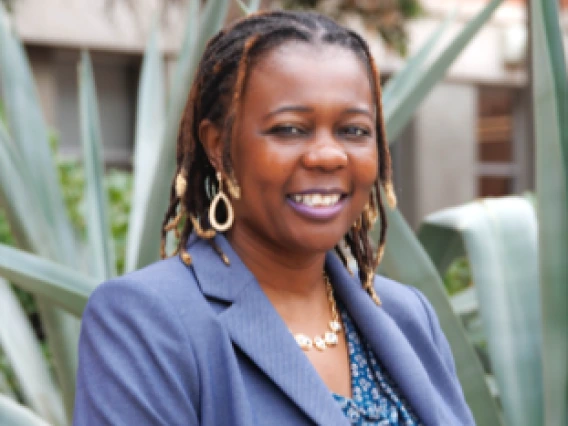 Photograph of a middle-aged black woman with blonde and brown hair, dreadlocks and dark eyes with a beautiful smile, wearing a purple jacket with a blue blouse and a gold necklace.