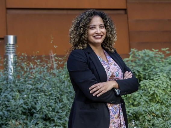 Portrait of Dr. Desiree Vega, a woman with curly brown hair, smiling and posing with arms crossed. She is wearing a black blazer over a floral-patterned dress. The background features green plants and a modern rust-colored metal wall.