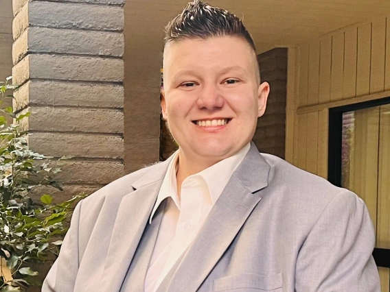 A person with short, neatly styled hair smiles at the camera while wearing a light gray suit and white dress shirt. The background features a modern outdoor setting with brick columns, plants, and a wooden-paneled wall.