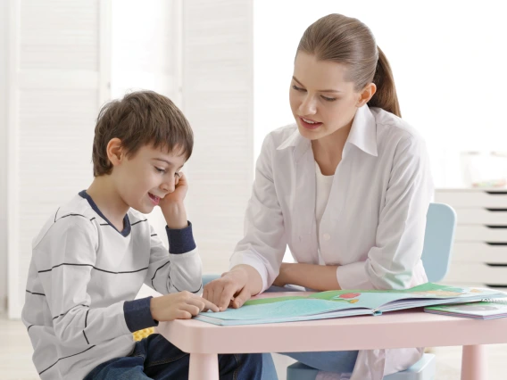 A young boy and a female educator sit at a small table, engaged in a learning activity. The educator, wearing a white coat, points to a colorful book as the boy smiles and follows along. The setting is a bright, modern classroom with white furniture and a small plant in the background.