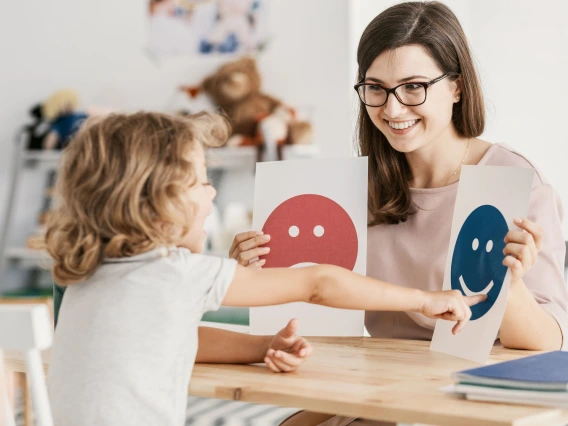 A smiling woman with glasses and a light pink blouse sits at a table holding two emotion cards, one with a sad face and one with a happy face. A young child with curly blonde hair excitedly points to the happy face. The setting is a bright, child-friendly classroom with toys and books in the background.