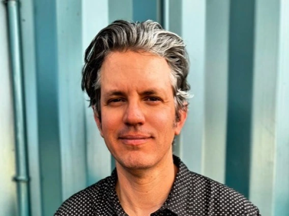 Portrait of Alex Lynch standing in front of a teal metal wall, wearing a black button-up shirt with white polka dots, and smiling slightly at the camera.