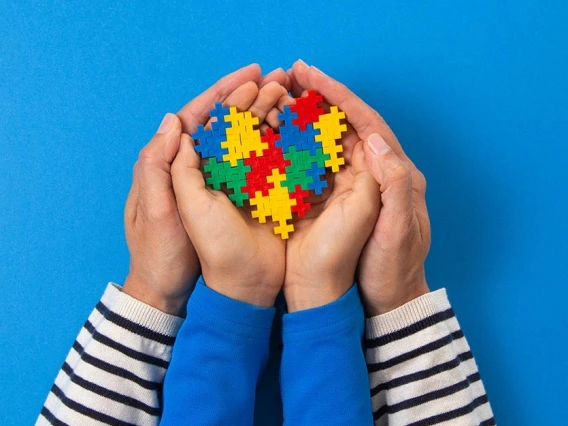 Adult and child hands gently holding a heart-shaped puzzle made of colorful interlocking pieces, symbolizing autism awareness, against a bright blue background.