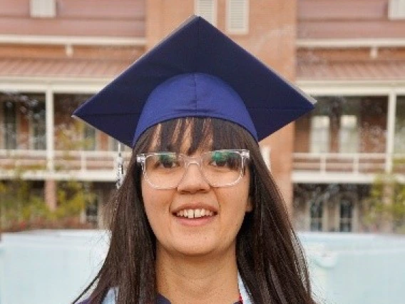 Graduating student Claire Adams stands in front of a university fountain wearing a navy blue cap and gown, light blue stole, and red and navy honor cords, smiling with long hair and glasses.