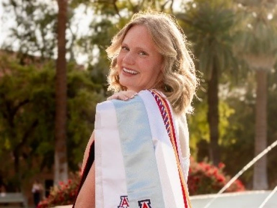 Graduating student Hadly Turner smiles over her shoulder while seated near a fountain, wearing multiple University of Arizona stoles and honor cords, including a Study Abroad stole and Arizona Ambassador sash.