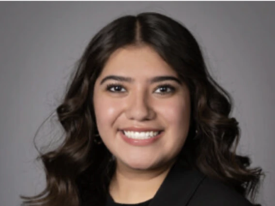 Professional portrait of a young woman with long wavy dark hair, wearing a black blazer with a Boys & Girls Clubs lapel pin, smiling in front of a neutral background.