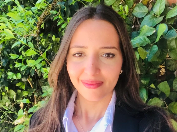 Yousra smiles in a professional headshot taken outdoors in front of green ivy. She wears a white collared shirt, a black blazer, and a delicate necklace, with long brown hair and natural makeup.