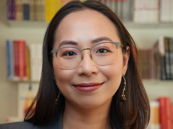 Professional portrait of Chi Nguyen smiling in front of a blurred background of colorful bookshelves. She wears glasses, dangling earrings, a delicate necklace with a round pendant, and a dark blazer over a light-colored top. Her hair is neatly styled, and she appears confident and approachable.