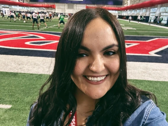 Frances María Montaño smiling on an indoor football field at the University of Arizona, with players practicing in the background. She is wearing a red UA lanyard and a name badge from the College of Education.
