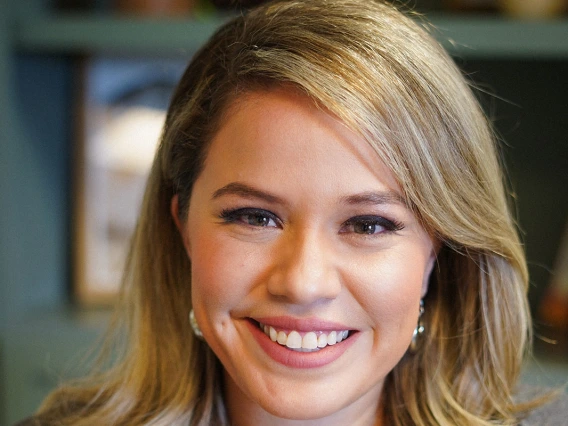 A warm, professional portrait of Laura Torres smiling directly at the camera. She has shoulder-length blonde hair styled to the side and is wearing a dark olive green top with a gray cardigan